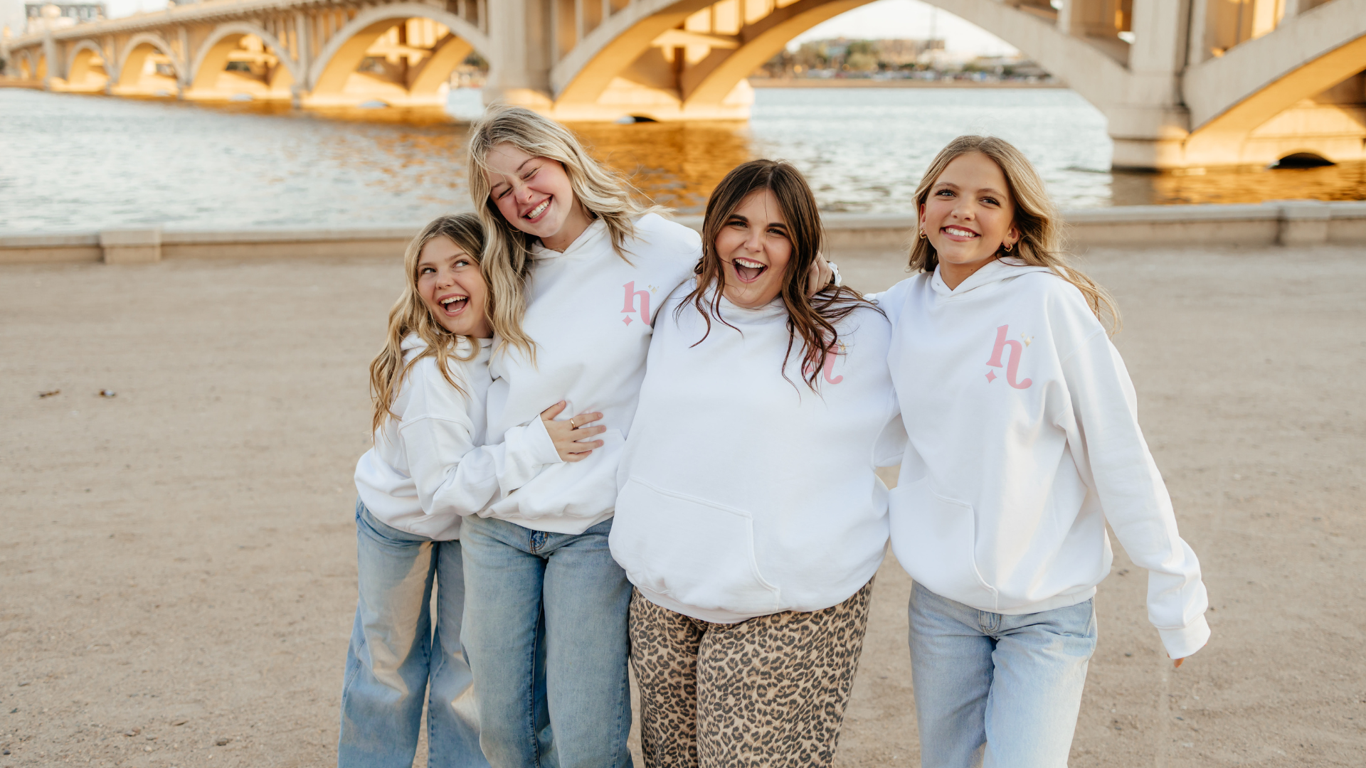 Four women posing together on a beach with a bridge in the background wearing The Headliner custom sweatshirts