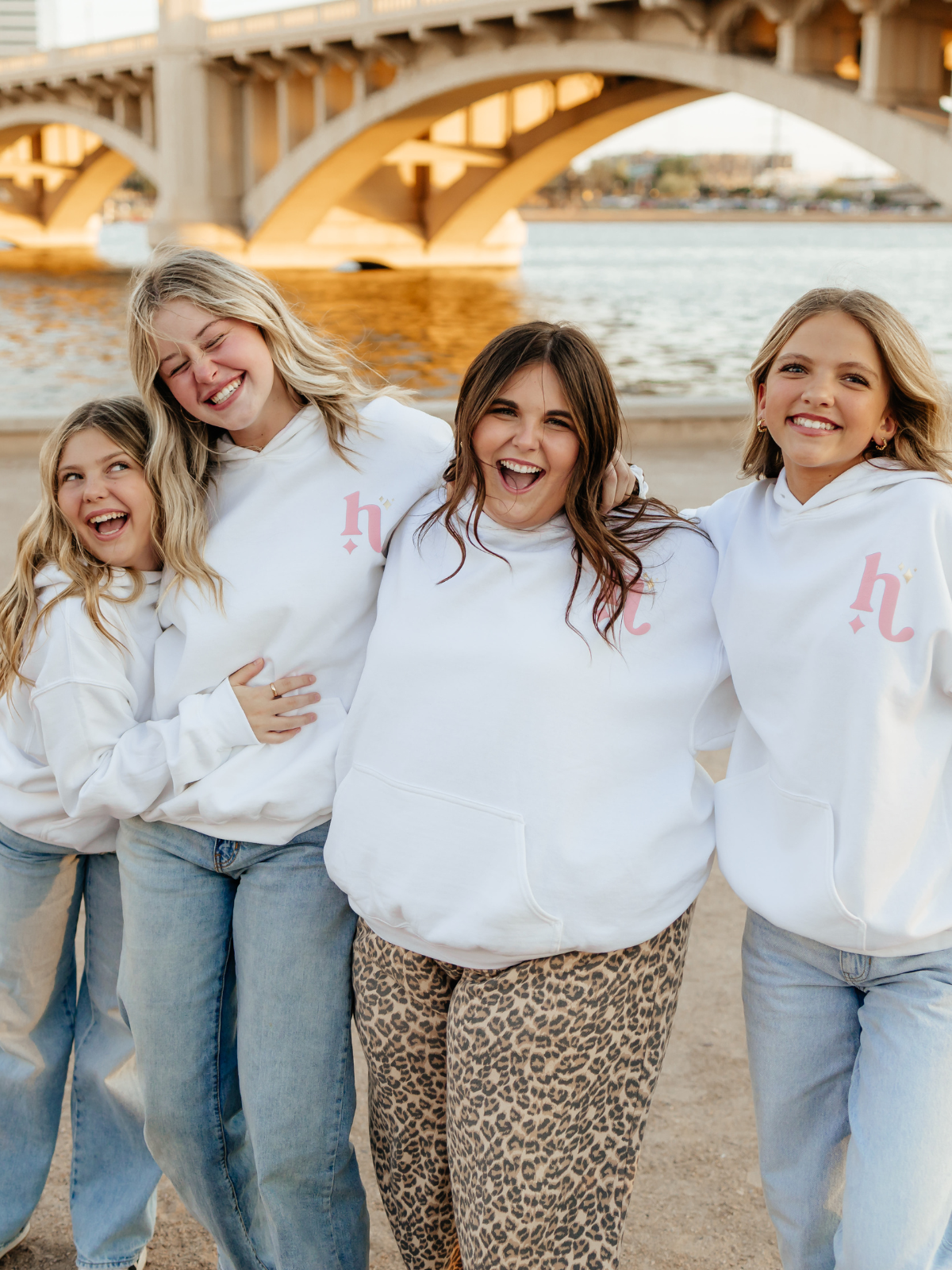 Four women posing together on a beach with a bridge in the background